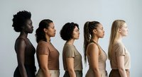 Five diverse women standing in profile line showing multicultural beauty and unity in neutral earth tone clothing against white studio background.