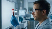Male scientist in safety glasses examining blood sample test tube in modern laboratory for medical research and diagnostic testing.