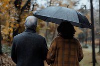 Elderly interracial couple sharing umbrella in autumn park, rear view of senior African American woman and Caucasian man walking together in rain.