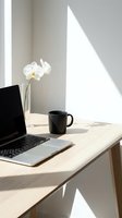 Minimalist workspace with laptop, black mug, and white orchid in glass vase on light wood desk, bathed in natural sunlight and geometric shadows for modern office or creative work concepts.