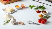 Fresh cooking ingredients arranged on white marble counter with herbs, spices, eggs, butter, flour, garlic, tomatoes and kitchen utensils for culinary preparation.