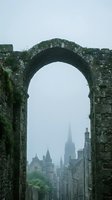 Ancient stone archway frames misty cityscape with historic spires and towers on overcast day creating atmospheric urban landscape view.