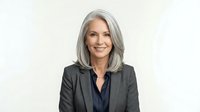 Mature woman with long gray hair wearing dark blazer and navy blouse, smiling at camera against white background. Corporate portrait for business, professional headshot, executive profile.