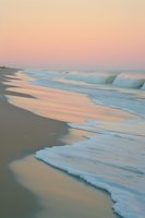 Gentle ocean waves rolling onto sandy beach during pink sunset, creating soft reflections and foam patterns along shoreline in pastel colors.