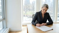 Middle-aged woman with blonde hair, Caucasian ethnicity, wearing professional black blazer, sitting at wooden desk writing on document near bright windows with urban cityscape view.