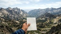 Caucasian male hand holding blank white card against mountain valley landscape with forested peaks, suitable for travel marketing, adventure tourism, and outdoor brand messaging.