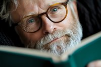 Senior man with gray beard and round eyeglasses reading a book, focused expression, suitable for education, literacy, and learning concepts.