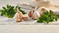 Fresh garlic bulbs and cloves with green parsley herbs on rustic white wooden table for cooking and culinary preparations in kitchen setting.