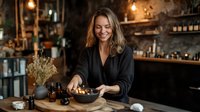 Young woman in black attire smiling while preparing food at rustic restaurant counter with warm ambient lighting and decorative dried flowers in background.