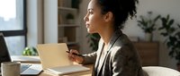 Young African American businesswoman working on laptop at home office desk with coffee cup and plants in modern workspace.