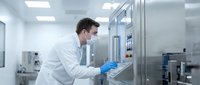Male scientist in white lab coat and blue gloves operating control panel in modern sterile laboratory facility for pharmaceutical research and development.