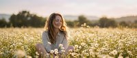 Young woman with long blonde hair sitting peacefully in blooming daisy field during golden hour sunset, enjoying nature and tranquil outdoor moment.
