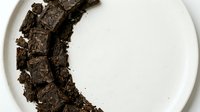 Crumbled dark chocolate pieces arranged in crescent moon shape on white plate, isolated on white background for food photography and culinary design projects.