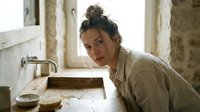 Young woman with dark hair in bun, wearing beige linen shirt, leaning over rustic sink in vintage bathroom setting, looking at camera with contemplative expression, natural daylight from window.