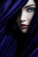 Mysterious partial face of a young woman with bright blue eyes and red lips emerging from dark shadows and deep blue fabric, dramatic lighting creates mystical mood.