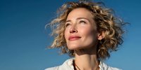 Young Caucasian woman with curly blonde hair looking up optimistically against clear blue sky background, expressing hope and positive mindset for wellness concepts.