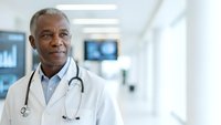 Confident African American male doctor in white coat with stethoscope standing in modern hospital corridor, professional healthcare worker portrait.