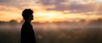 Young African American man silhouette standing in misty field during golden hour sunrise with dramatic cloudy sky and warm sunlight rays creating peaceful morning atmosphere.