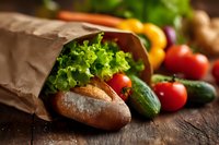Fresh organic vegetables and artisan bread spilling from brown paper bag on rustic wooden table. Healthy grocery shopping and farm-to-table cooking concept.