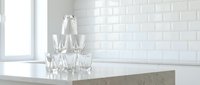 Young woman with fair complexion and blonde hair, wearing professional business attire, standing in a modern kitchen with white subway tile backsplash, concrete countertop, and clear glass drinking glasses arranged on counter near window.