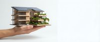 Hand holding miniature modern eco-friendly house model with solar panels and green plants on white background for sustainable architecture concepts.