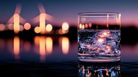Glass of sparkling water with ice cubes against blurred city lights background at sunset, creating atmospheric bokeh effect and reflections on glass surface.