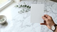 Cropped hand holding blank white card on marble surface with eucalyptus leaves and accessories for stationery mockup and branding projects.