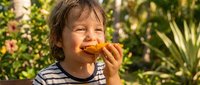 Young boy with dark hair, Caucasian ethnicity, eating a croissant outdoors in a sunny garden setting with green foliage and flowers in the background.