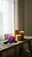 Three beeswax pillar candles on a rustic wooden table with a pink orchid and black zen stones near a soft linen curtain window.