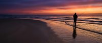 Solitary figure standing on beach at sunset with dramatic orange and purple sky reflecting on wet sand and gentle waves washing ashore creating peaceful moment.