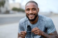 Smiling athletic Black man in gray workout shirt tying running shoes outdoors, ready for morning jog or fitness training session.