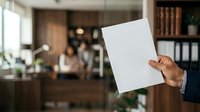 Businessman holding blank white paper sheet in modern office with colleagues working in background, suitable for corporate communications and business presentations.