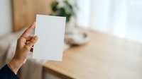 Hand holding blank white paper sheet in modern office workspace with wooden desk and blurred background for mockup and branding purposes.