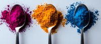 Vibrant spices and pigment powders in pink, orange, and blue colors displayed on silver spoons against white background, scattered powder creates artistic effect.