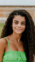 Young woman with long dark curly hair and olive complexion wearing bright green strapless top, smiling at camera outdoors with brick wall background. Portrait for lifestyle and beauty content.
