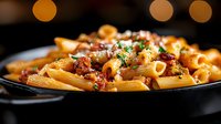 Penne pasta with crispy bacon bits and parmesan cheese garnished with fresh herbs in black plate against warm bokeh background, close up view of Italian cuisine.
