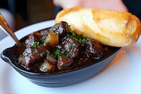 Tender braised beef chunks in rich dark gravy sauce with potatoes and fresh herbs, served in black ceramic dish with crusty French bread for restaurant menu.