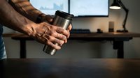 Professional businessman holding stainless steel travel mug in modern office workspace with computer monitor and city view background.