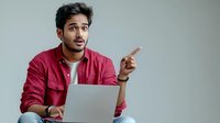 Young South Asian man in red shirt pointing sideways with surprised expression while working on laptop against neutral background.