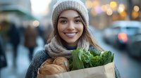 Young Asian woman in winter hat and scarf holding paper grocery bag with fresh produce and bread, smiling outdoors with blurred city lights background.