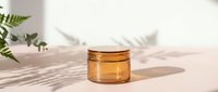 Amber glass cosmetic jar with golden lid on neutral background with natural fern shadows, minimalist skincare product photography for beauty branding.