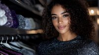 Young African American woman with natural curly hair smiling in retail store, wearing sparkly evening dress. Soft bokeh lighting creates elegant shopping atmosphere.