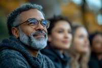 Mature African American man with gray beard and glasses smiling confidently outdoors, wearing casual sweater, with diverse group of people in soft focus background.
