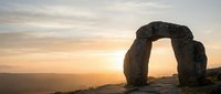 Ancient stone arch monument silhouetted against golden sunset sky, featuring weathered megalithic structure framing distant landscape for travel and historical content.