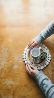 Woman hands holding wicker tray with white cup, small succulent plant and decorative stones on wooden table. Cozy home decor and mindful living concept.