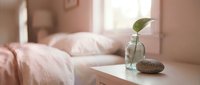 Cozy bedroom interior with soft pink tones, a glass vase with a green leaf, and a stone on a white nightstand in warm light.
