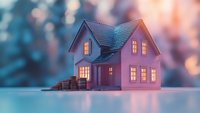 Miniature house model with glowing windows and stacked coins against dreamy bokeh background in pink and blue tones, representing real estate investment and savings.