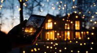 Smartphone capturing glowing house with Christmas lights at dusk, warm bokeh lights create magical holiday atmosphere against winter evening sky.