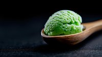 Green matcha ice cream scoop in wooden spoon on dark background, selective focus with soft bokeh effect, showcasing creamy texture and vibrant color.