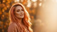 Young Caucasian woman with long wavy red hair smiling warmly in golden autumn sunlight with bokeh background for lifestyle and beauty content.